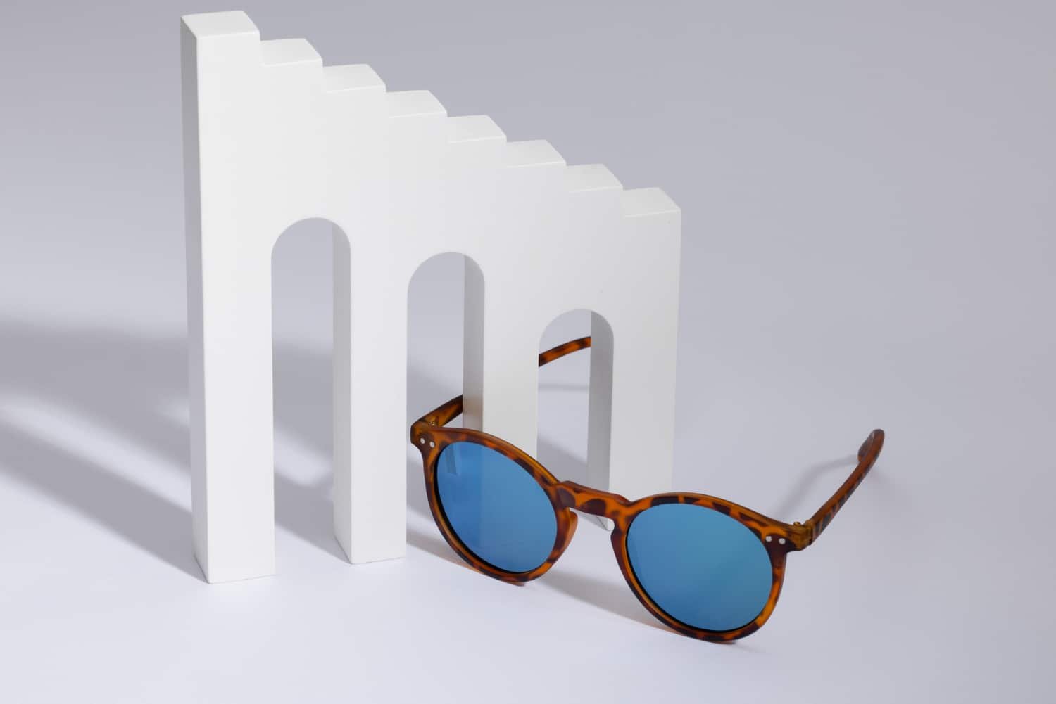 Lunettes de soleil avec monture écaille et verres bleus posées à côté d'un décor blanc abstrait.