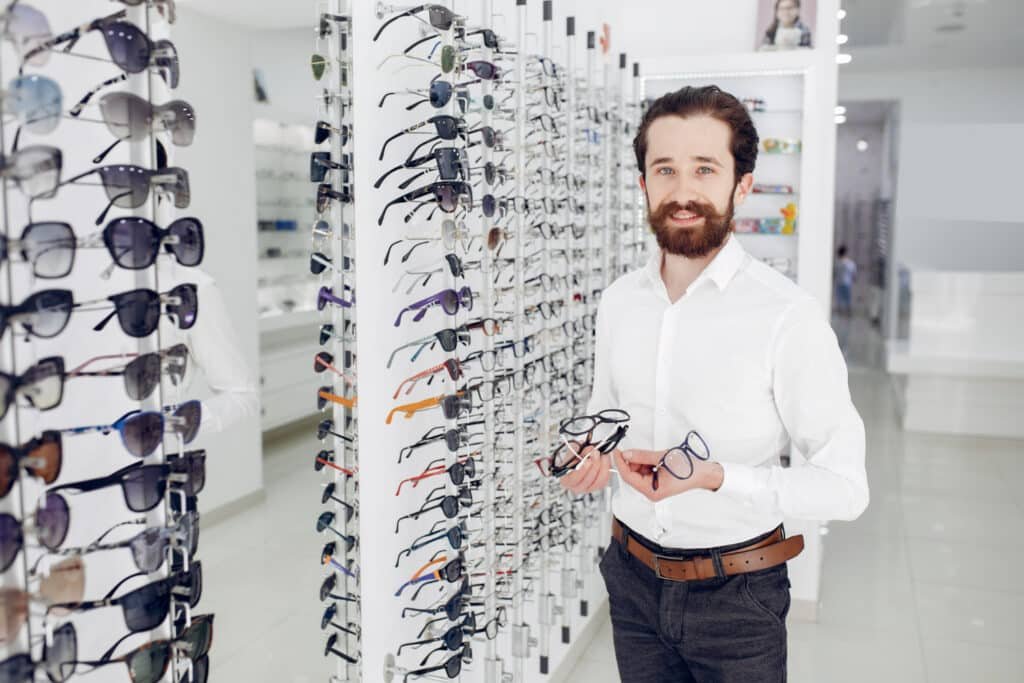 Opticien barbu tenant des lunettes dans un magasin bien approvisionné en lunettes de soleil et de vue.